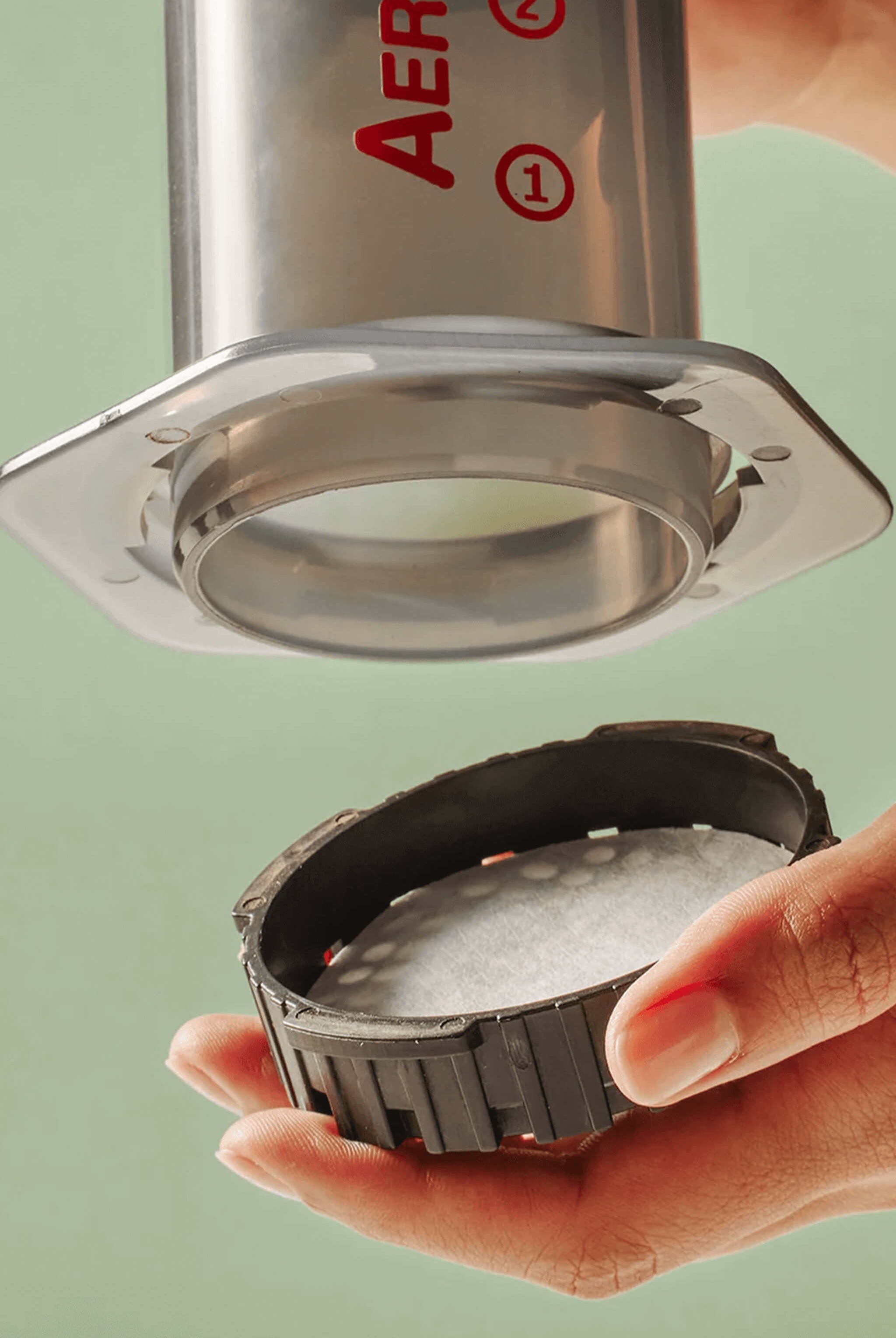 Hand holding a metal filter with 'Aeropress' branding against a light green background