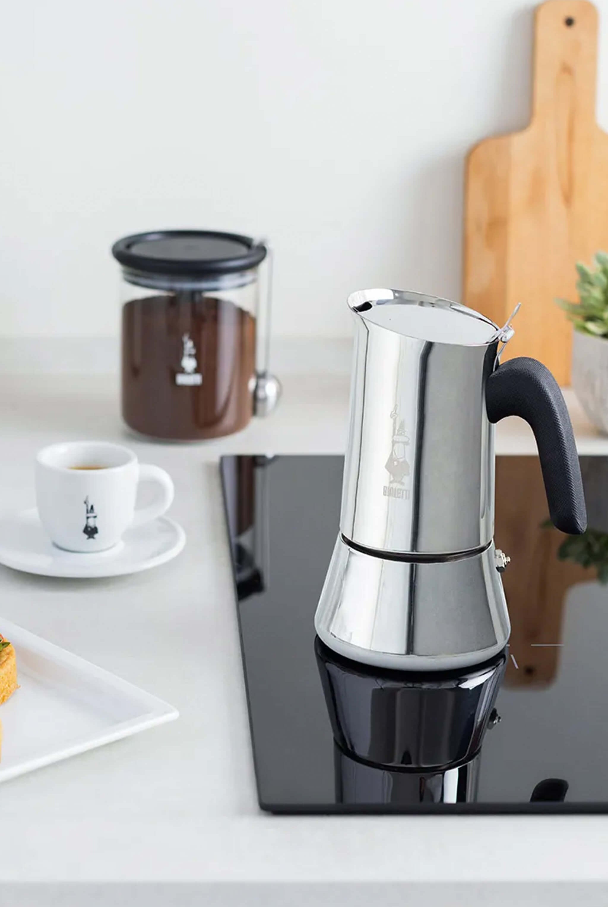 Silver espresso maker on a kitchen counter with a cup of coffee and a jar of coffee beans in the background.