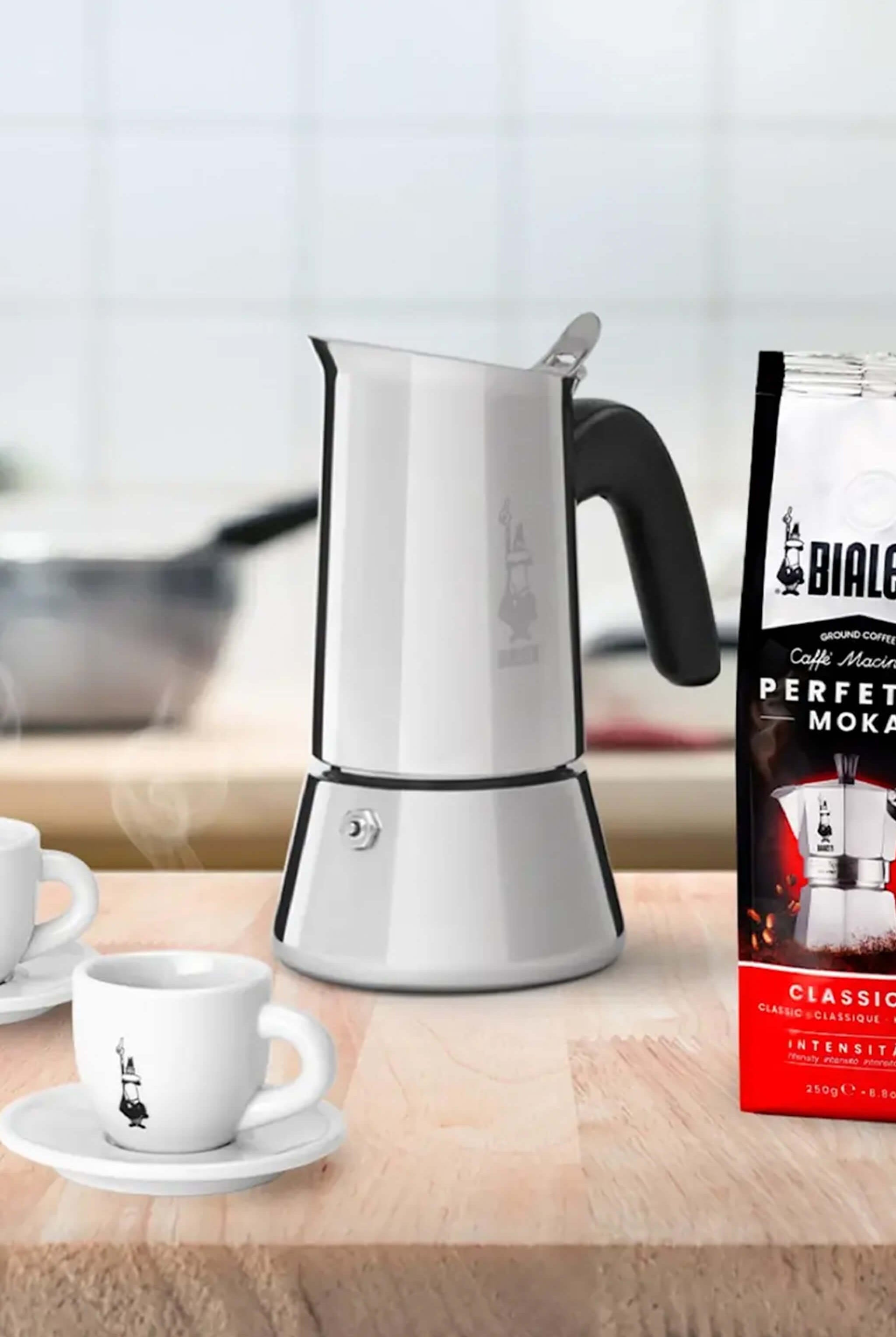 Stainless steel espresso maker with a cup and Bialetti coffee beans on a wooden surface.