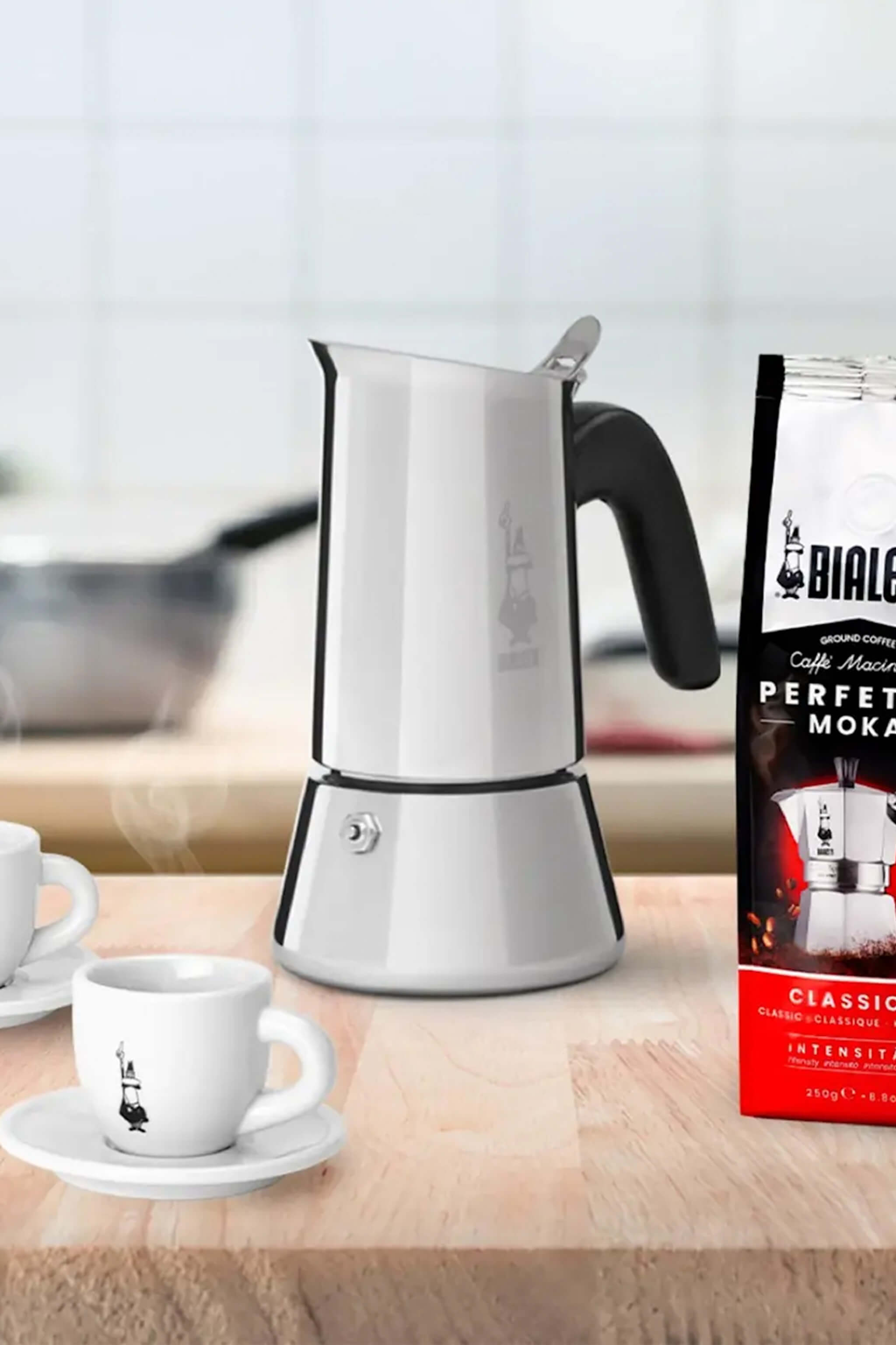 Stainless steel espresso maker with a cup and Bialetti coffee beans on a wooden surface.