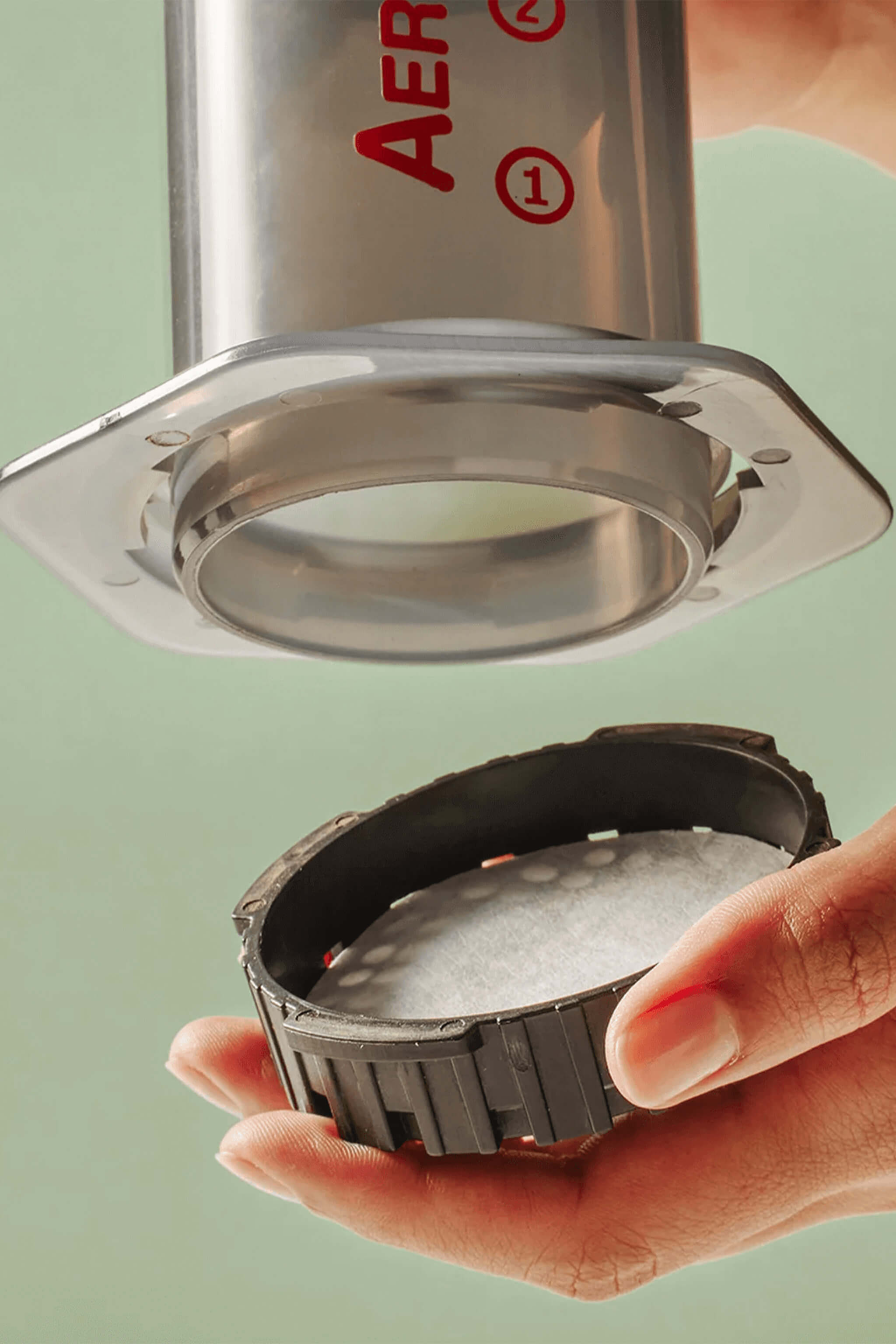 Hand holding a metal filter with 'Aeropress' branding against a light green background