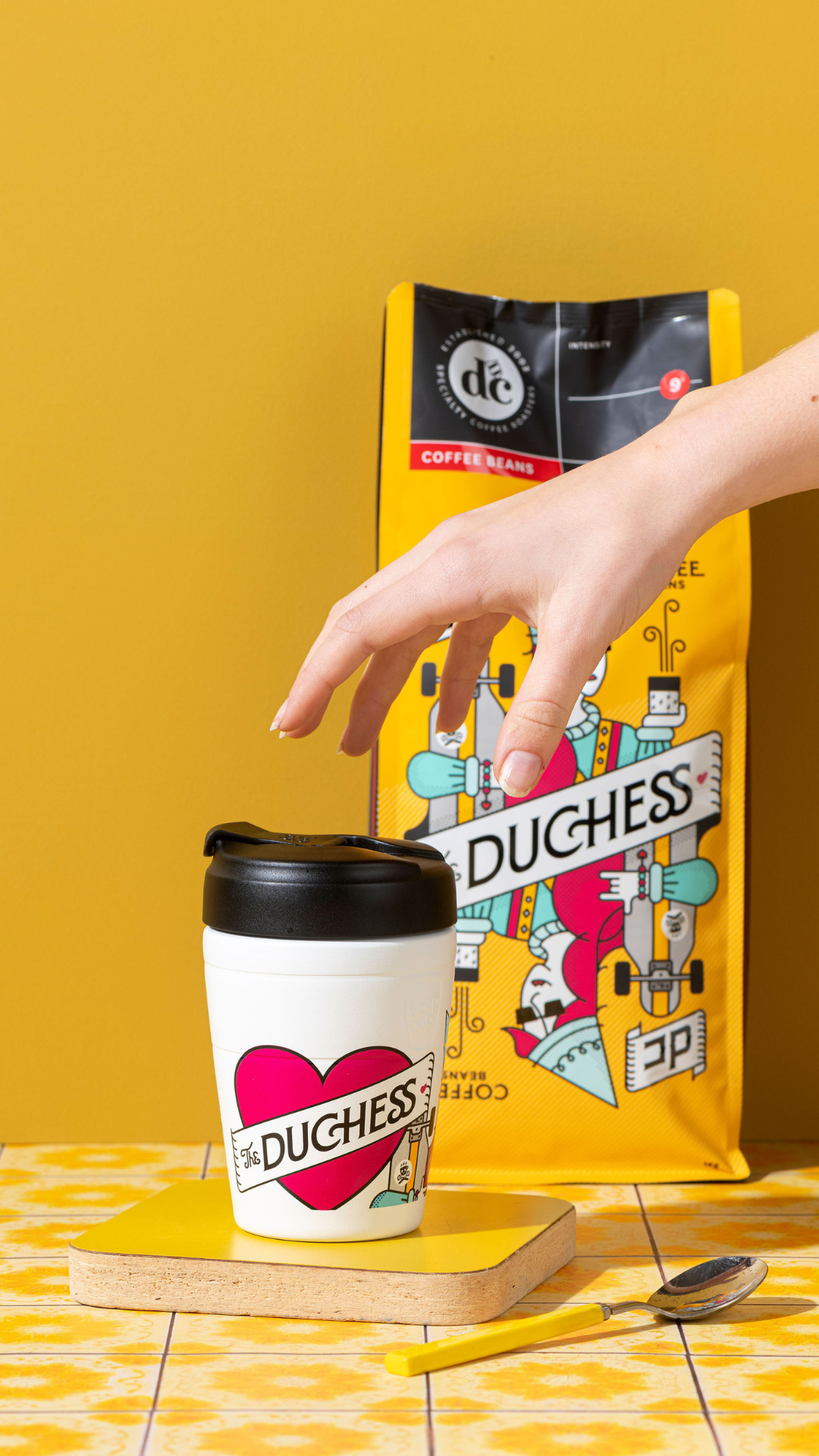 Hand reaching for a Duchess KeepCup coffee cup with coffee beans on a yellow background